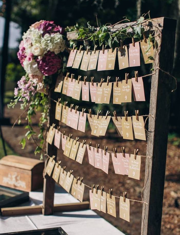 plan de table mariage en grand cadre rustique en bois avec des étiquettes noms invités et nombre table en papier coloré, guirlande verte et bouquet de fleurs colorées
