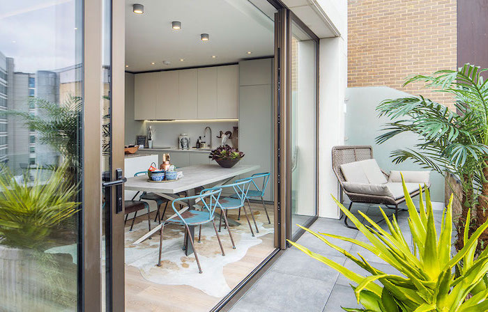 ouverture cuisine sur une terrasse revêtue de béton, avec palmiers, plantes succulentes, chaise en rotin, coussins gris
