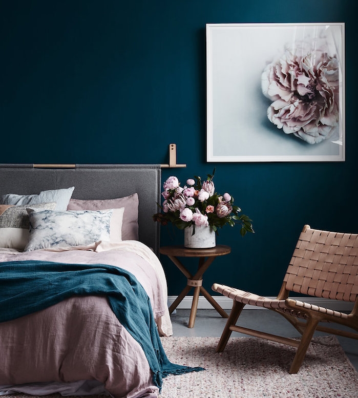 peinture chambre adulte couleur bleu foncé, linge de lit rose, bleu et blanc, fauteuil, table basse en cuivre, avec bouquets de fleurs dans in vase, cadre peinture fleur