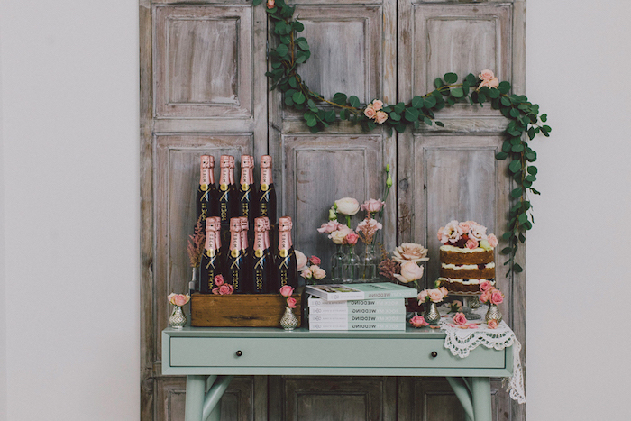 deco mariage vintage, fond de portes en bois brut avec decoration de guirlande fleurie, table vert pastel avec des bouteilles de champagne, gateau, bouquins et decoration de fleurs