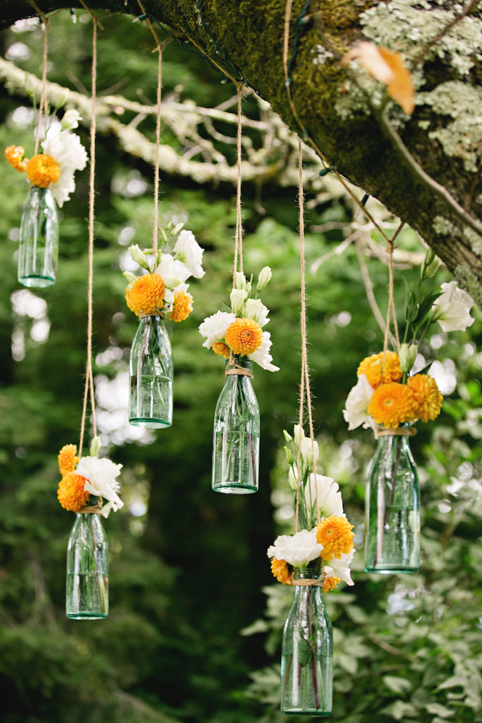 exemple de deco mariage pas cher, des bouteilles en verre avec des fleurs orange et blancs dedans, petits vases suspendus par des ficelles à une branche d arbre