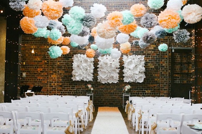 deco salle mariage, des fleurs en papier de soie colorées, couleur orange, bleu, blanc et gris, chaises blanches, mur en briques décorations blanches en papier