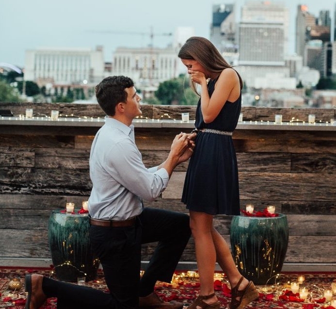 demande en mariage romantique au toit d un bâtiment, femme en robe noire, homme cravate pantalon, sol recouvert de pétales de roses, bougies et guirlande lumineuse