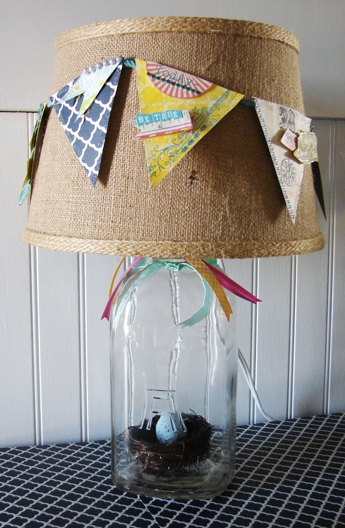 une lampe à poser avec abat-jour décoré de fanions et support bocal contentant un nid doiseau décoratif