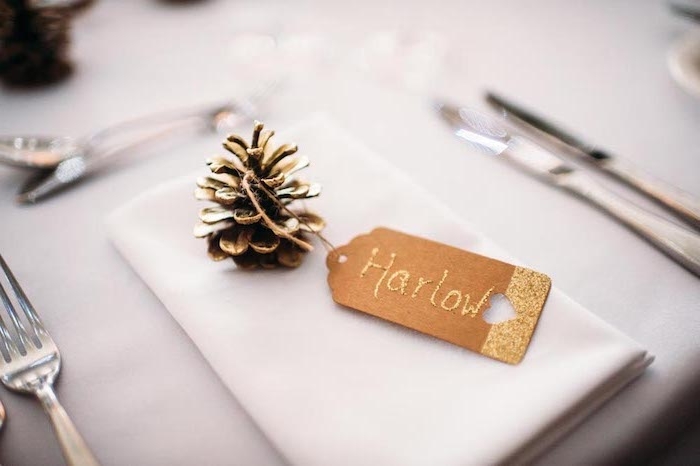 deco mariage pas cher, pomme de pin repeinte de couleur or avec étiquette en papier kraft doré sur une serviette blanche