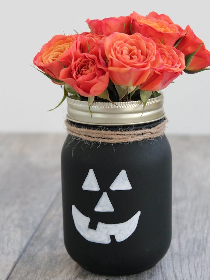 pot de fleur, repeint en noir avec des traits de visage blancs, bricolage halloween avec pot en verre et bouquet de roses dedans