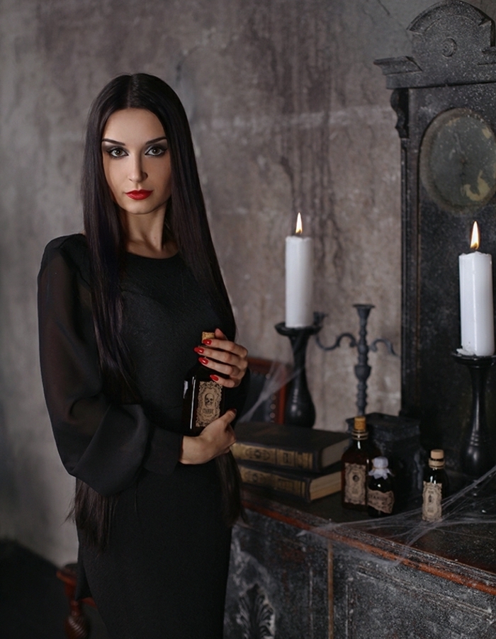 Idée tenue morticia addams, robe noire avec manches transparentes, livres grimoires, bougies fioles sur le fond, cheveux lisses longues teinte pale