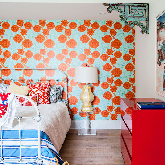 modele de papier peint vintage avec imprimé de fleurs orange sur un fond bleu, linge de lit blanc et bleu et coussins blanc, rouge et bleu, parquet bois clair, commode rouge