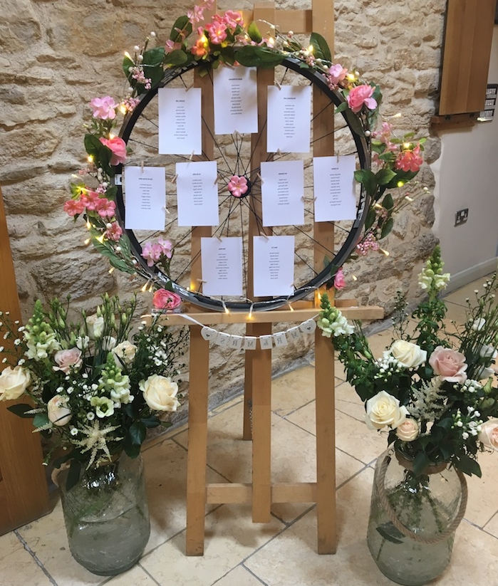 plan de table mariage original, style campagne chic, roue de bicyclette avec des étiquettes avec les noms des invités sur un chevalet et dame jeannes en verre, deco florale