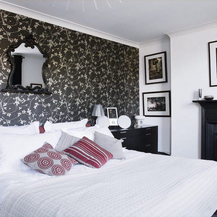 idée de tapisserie chambre sur un seul pan de mur, motif fleurs sur un fond noir, inge de lit blanc, coussins gris et rouge, commode noire, miroir vintage