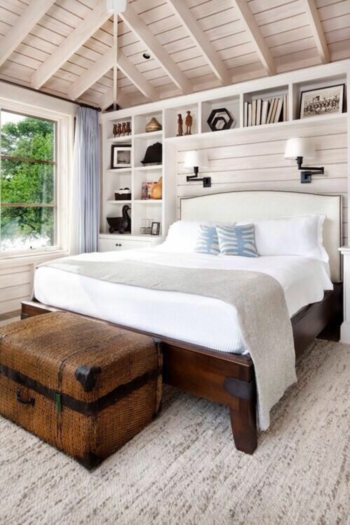 aménager une petite chambre sous toit en pente, avec grand lit en bois marron foncé, grande malle marron au pied du lit, étagères rangement au dessus du lit 