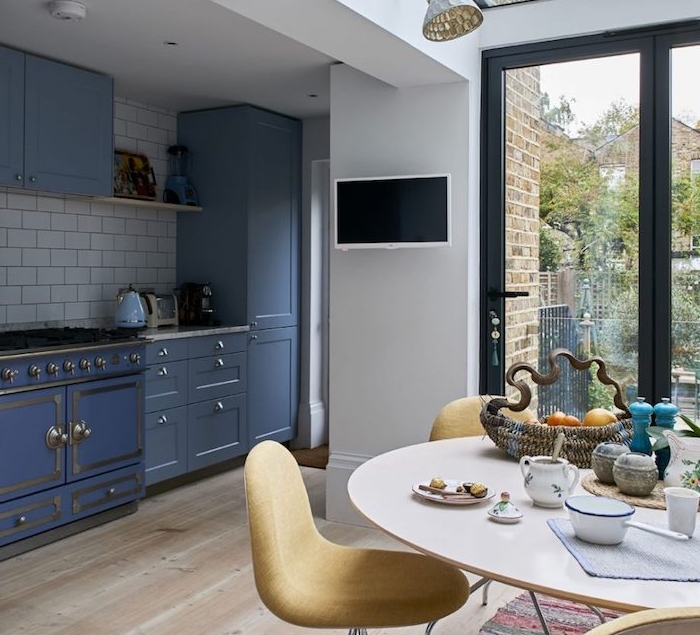 amenagement petite cuisine bleue avec credence en carrelage blanc, parquet clair, ouverture sur salle à manger table ronde et chaises jaunes