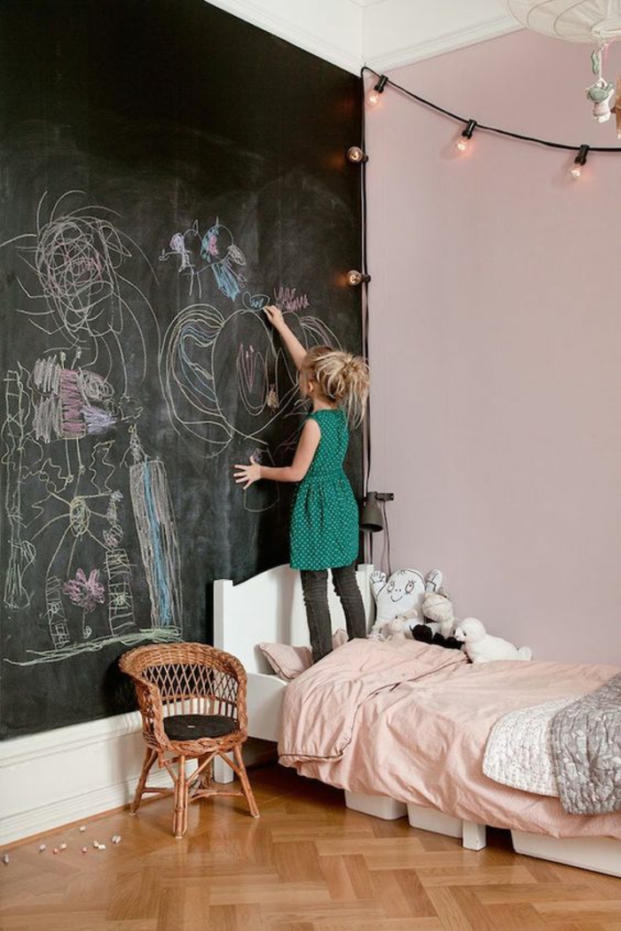 décoration chambre adulte avec tableau noir sur un mur entier pour dessiner et pour écrire, décoration murale avec des guirlandes lumineuses en forme d'ampoules , lit bas en couleur blanche, avec petit fauteuil en canne tressée à côté du lit