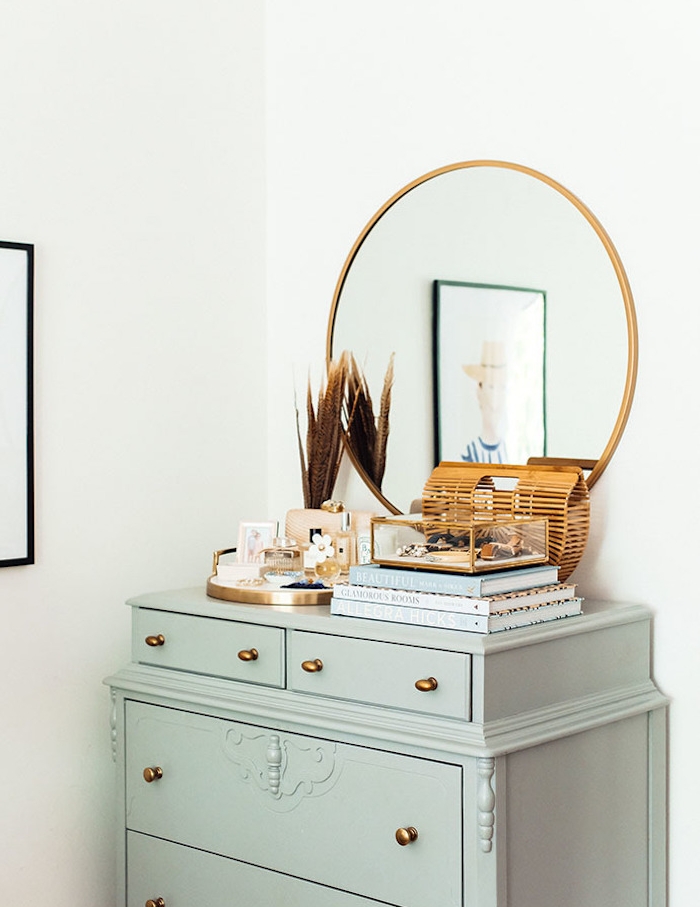 commode vintage et coiffeuse vert gris céladon, pile de livres, miroir rond dans cadre doré, produits de toilette, mur blanc