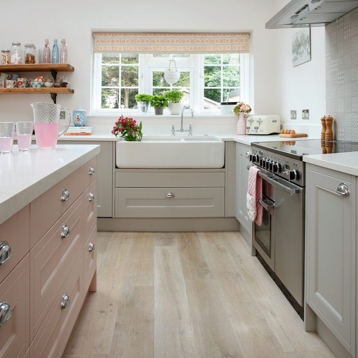 cuisine ancienne avec facade gris clair, plan de travail et évier blanc, ilot central rose clair, parquet bois clair, étagères en bois et metal