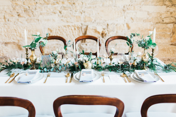 deco table mariage, nappe blanche, centre de table feuillages, fleurs dans de petits vases dorées, bougies blanches, mur de fond en pierre, réception dans une grange