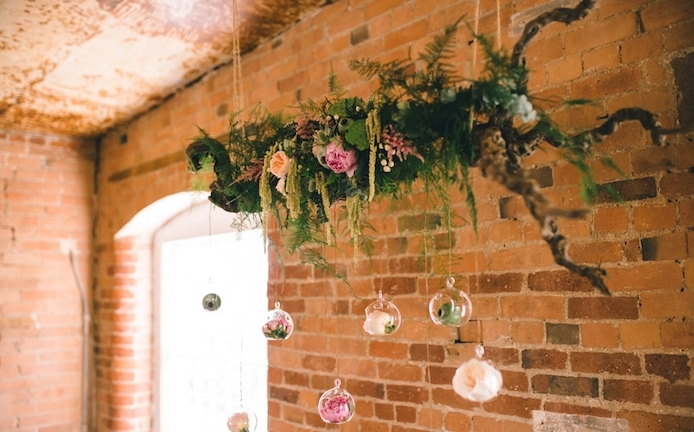 mariage champetre chic, branche de bois décoré de feuillages et fleurs avec de petits terrariums suspendus avec des fleurs dedans, fond de murs en briques