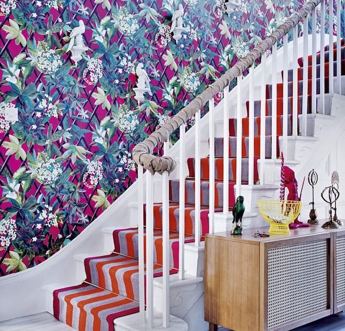 déco montée d escalier avec mur habillé de papier peint à motifs floraux sur un fond fuchsia, habillage escalier de tapis à rayures, orange, rouge, gris et rose, main courante avec tissu gris enveloppé autour 