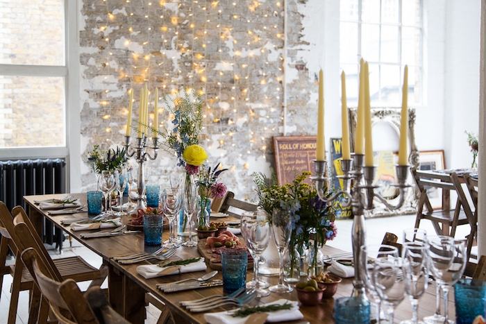 decoration mariage champetre, table et chaises en bois, verres bleues, bouteilles en verre avec des fleurs, bougeoir argenté avec bougies jaunes, guirlande lumineuse sur un mur usé
