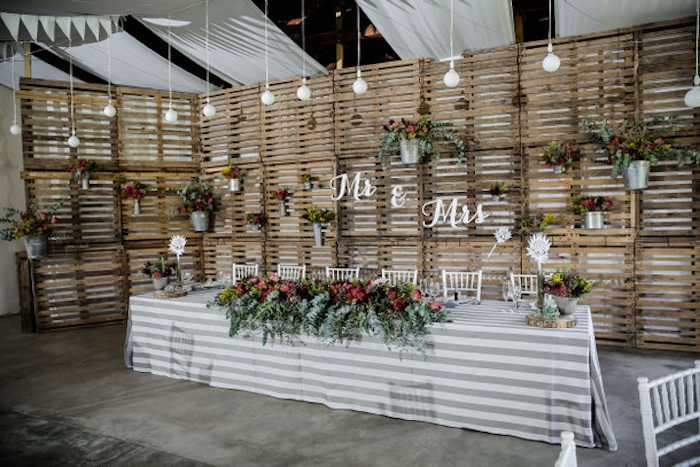 un mur en palettes de bois rustique avec seaux et boites de conserve fleuries, table avec nappe blanche et grise, decoration florale