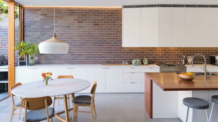 modele de cuisine ouverte blanche, avec crédence en briques, ilot cental avec plan de travail en bois, salle à manger table ronde et chaises en bois et tissu
