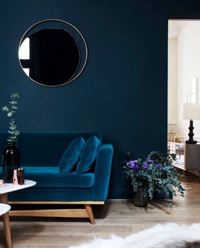 idée de canapé bleu pétrole adossé à un mur bleu marine avec un miroir rond accroché, parquet clair, table basse en bois, tapis peau animal