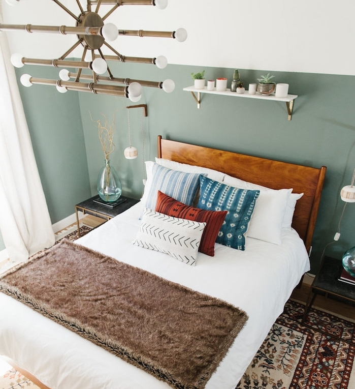 chambre design aux murs couleur vert de gris, en guise de tete de lit, lustre original, lit en bois avec linge de lit blanc, coussins en bleu, rouge et blanc, tapis oriental, étagère blanche rangement petites plantes