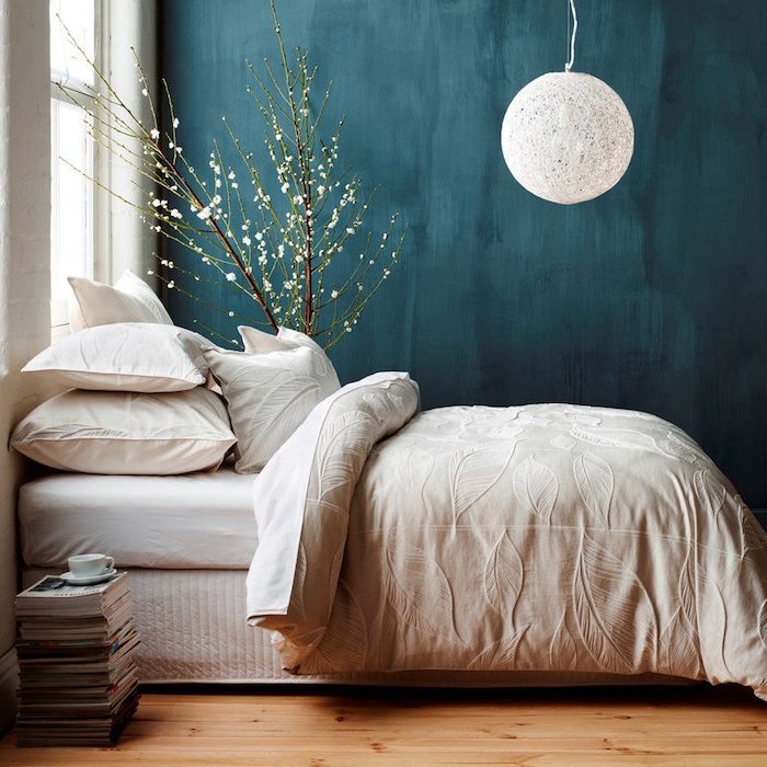 chambre à coucher couleur bleu pétrole en peinture murale couleur foncée, suspension boule blanche, linge de lit blanc, parquet clair, pile de magazines, arbre decoratifs