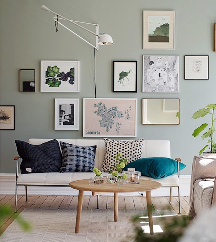 mur repeinte de couleur vert pastel, nuance céladon dans un salon design avec canapé ivoire, décoré de coussins bleu, gris et noir, table basse en bois, tapis gris clair, mur de cadres decoratifs, plantes vertes