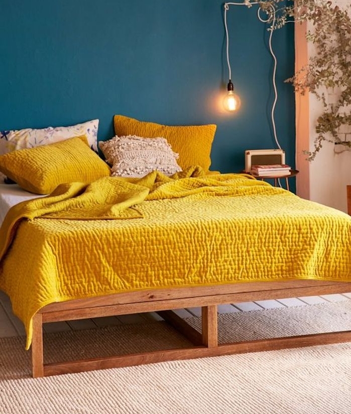 un pan de mur nuance de bleu, couleur pétrole, lit en bois, linge de lit blanc et jaune, lampe de nuit ampoule électrique, tapis beige, plante verte