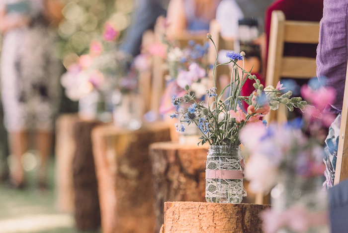 deco champetre cérémonie mariage en bûches en bois décorés de pots en verre, décorés de dentelle et ruban rose et fleurs champetre