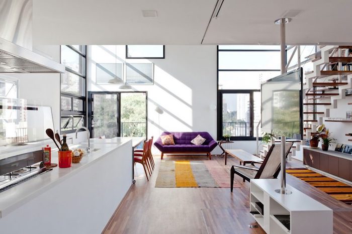 modele de cuisine americaine blanche ouverte sur salle à manger avec table blanche et chaises en bois, tapis à rayures et canapé violet, escalier en bois