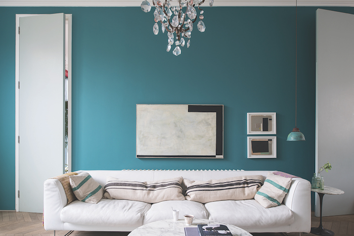 modele de salon bleu petrole, mur repeint en bleu de tonalité pastel, canapé blanc, décoré de coussins beife à rayures vertes et marron, table basse en marbre, lustre élégant