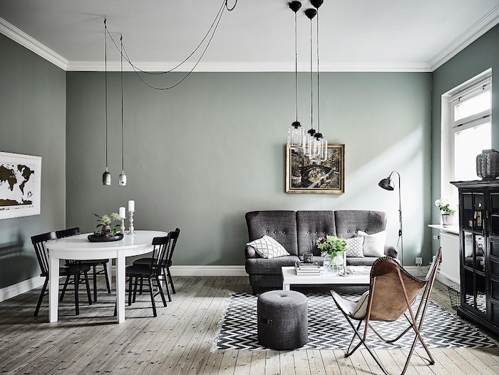 murs peinture vert de gris céladon dans un salon salle à manger scandinave, parquet en bois clair, suspensions originales, canapé et tabouret gris, tapis chevron noir et blanc, chaise en cuir, table blanche et chaises noires