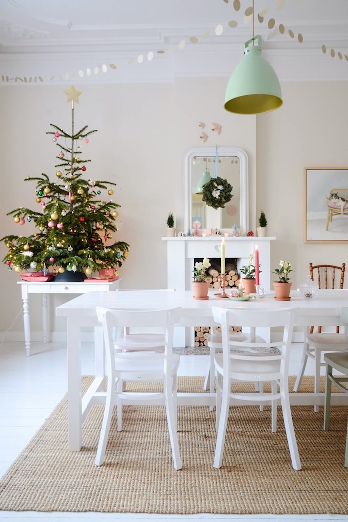 petit sapin de noel naturel décoré de boules de noel rouges, dorées, roses et blanches dans une salle à manger vintage scandinave, cheminée, chaises et table blanches