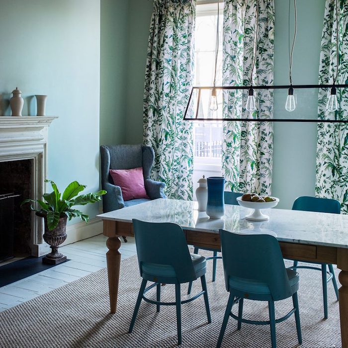 modele de salle à manger vert pastel, teinte céladon, table en bois avec plateau en marbre, chaises bleu canard, tapis gris, cheminée rustique, linge de lit blanc et vert, parquet clair blanchi, lustre en ampoules électriques
