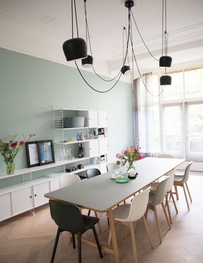 modele de salle à manger vert celadon, parquet clair, table en bois avec plateau gris, chaises scandinaves blanches et une chaise noire, suspensions noires, meuble blanc avec plusieurs étagères