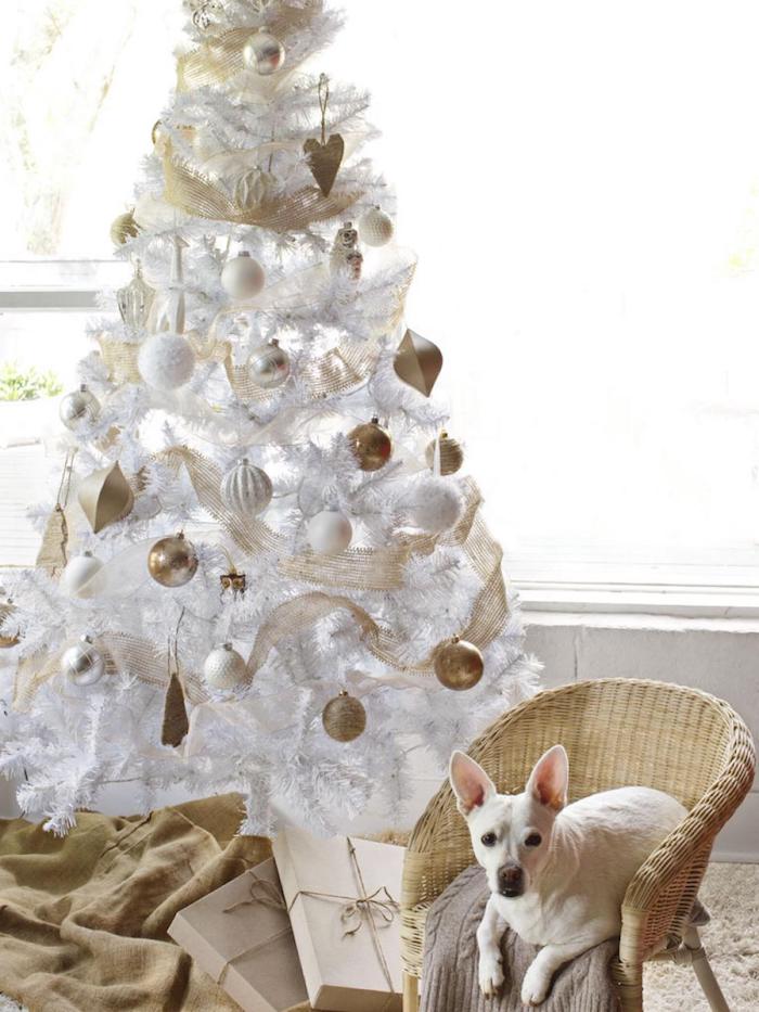 sapin de noel blanc décoré de boules de noel blanches et or, petite chaise en rotin, paquets cadeaux en papier kraft, couverture beige