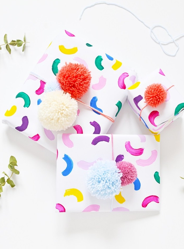 comment faire un paquet cadeau en papier blanc à motif arc en ciel coloré et décoration de pompons en rouge, bleu et blanc