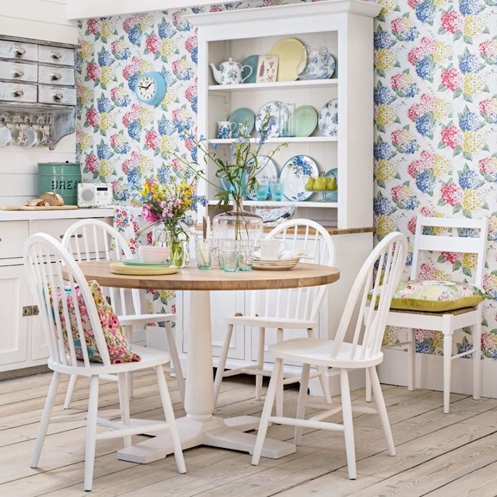 modele de cuisine campagnarde, style shabby chic avec mur habillé de papier peint à imprimé floral, table et chaises coin repas en bois, facade cuisine blanc et vaisselier blanc, décorée de vaisselle fleurie