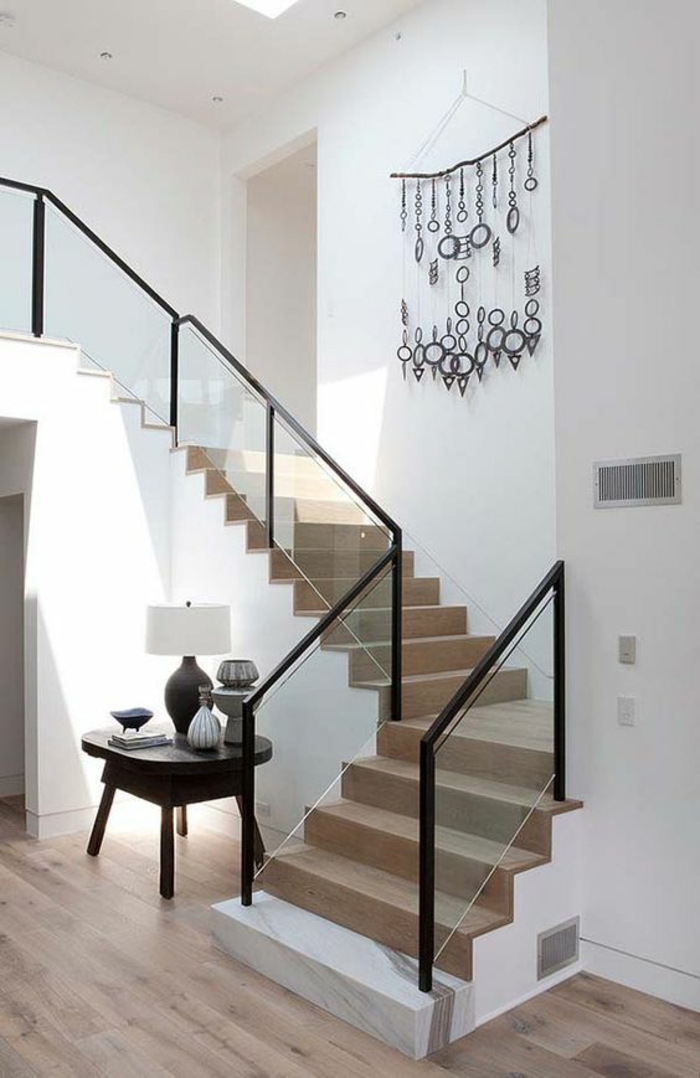 escalier flin en bois clair avec base blanche, garde corps en métal noir et verre blanc transparent, petite table carrée noire avec lampadaire avec abat-jour blanc, murs tout blancs 