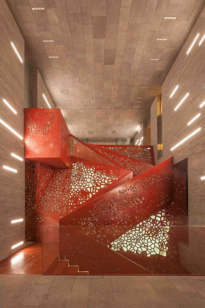 escalier design avec garde corps en couleur rouge, décoré de motifs ajourés, marches qui montent et qui descendent, murs et plafond en couleur beige nuances claires et foncées 