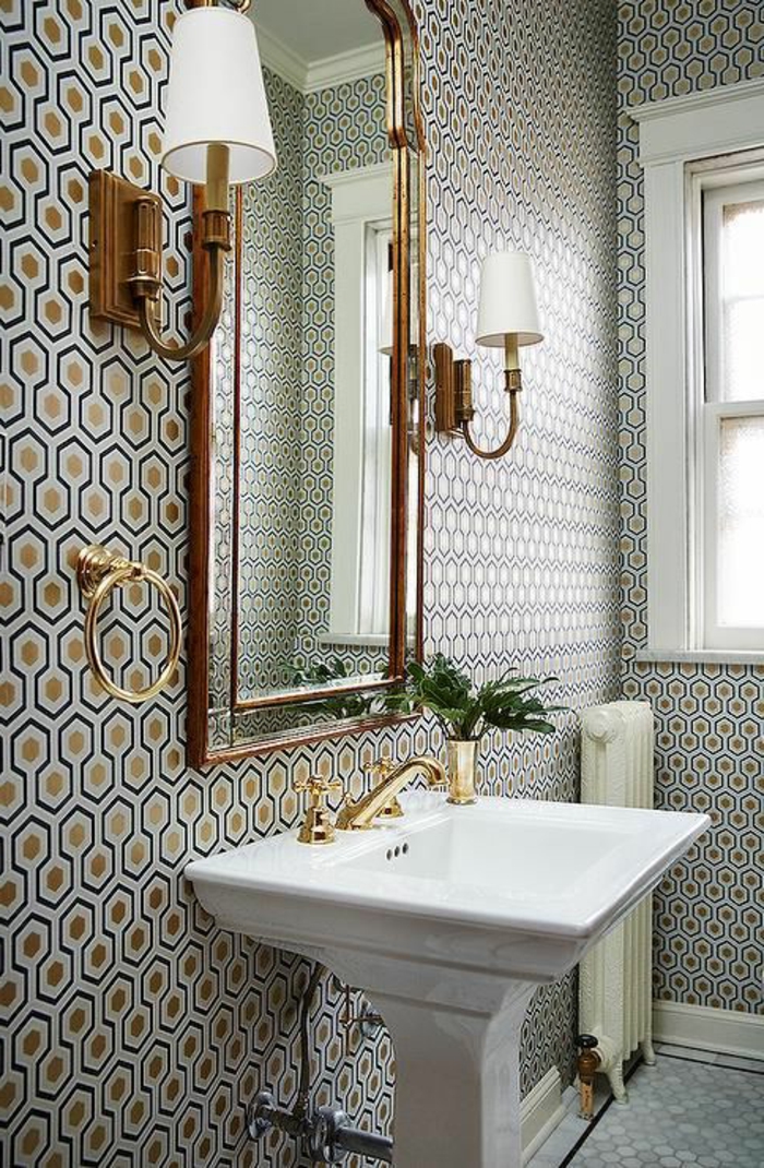 papier peint liberty en couleur bleu marine et or, aux motifs ruches, dans une salle de bains en style classique, avec grand lavabo blanc en style vintage, deux lampadaires appliques muraux avec des abat-jours blancs 