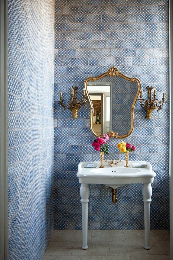 papier peint trompe l'oeil dans une salle de bains en style classique, avec miroir en forme baroque au cadre en métal couleur or, avec deux appliques murales en forme de bougeoirs, en style château, mur en bleu et blanc, effet de mosaïque à l'ancienne 