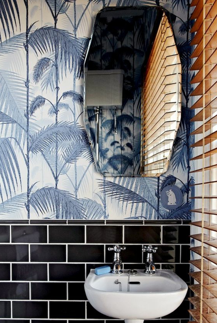 papier peint liberty avec des grandes feuilles de palmier en couleur bleue, miroir en forme géométrique, moitié du mur en briques noires, petit lavabo blanc à deux robinets, stores en beige