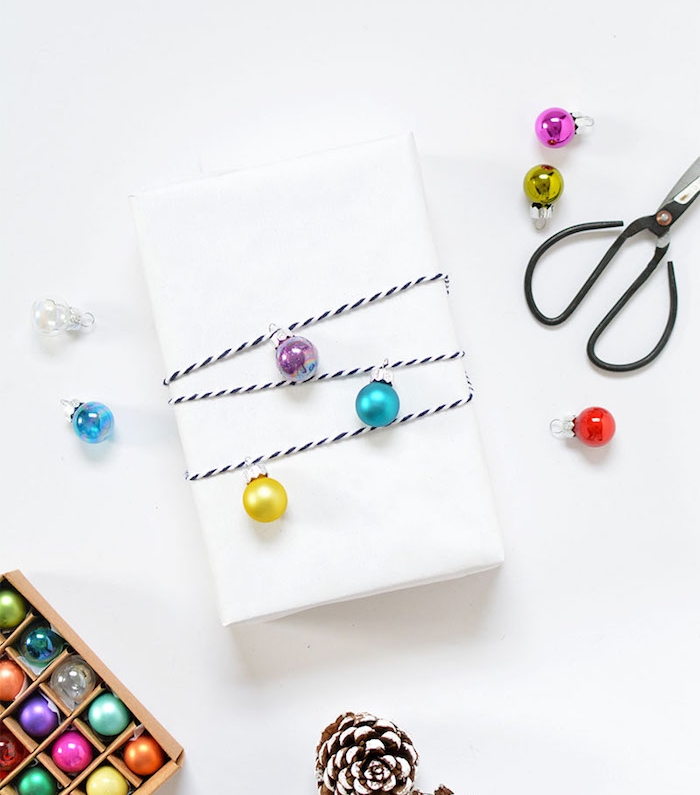 comment faire un emballage cadeau en papier blanc, décoré de petites boules de noel colorés sur une ficelle noir et blanc