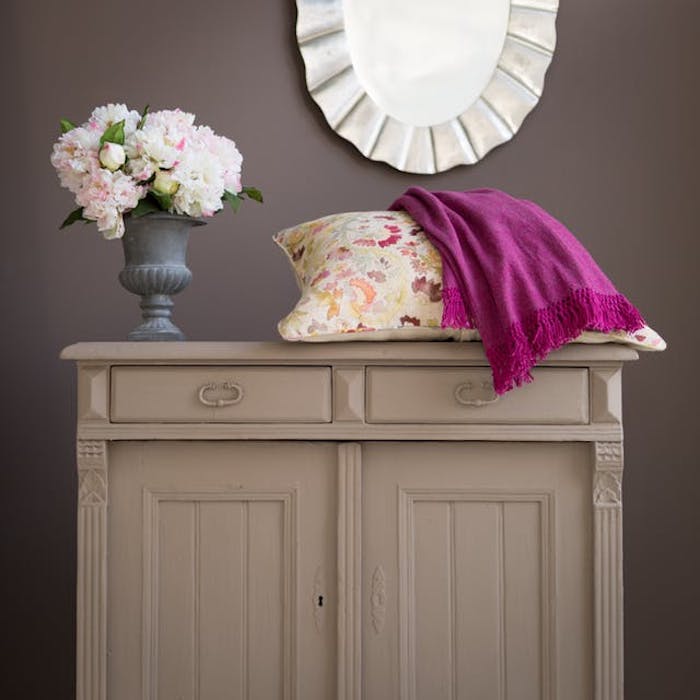 meuble relooké, commode repeinte de couleur grise, couverture mauve et coussin coloré à imprimé floral, bouquet de fleurs, mur gris foncé, miroir rond