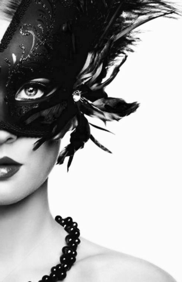 photo femme noir et blanc, avec masque vénitien aux grandes plumes noires et collier de perles noires, bouche charnue, look mystérieux