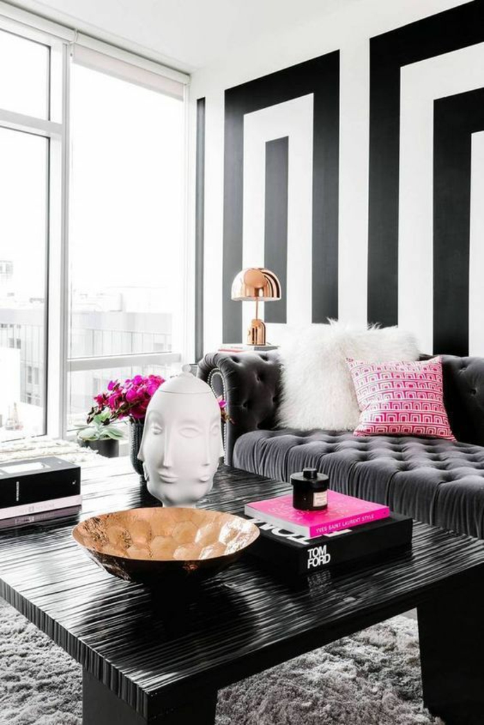 salon moderne de luxe avec canapé en gris anthracite, murs aux motifs graphiques en noir et blanc, table carrée en matière PVC noire, tapis en gris pastel, objet déco visage aux multiples faces 