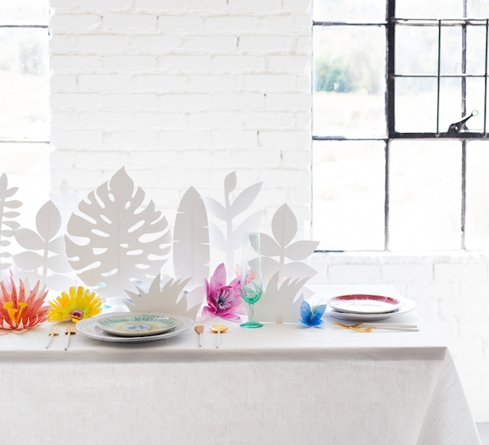 centre de table de feuilles tropicales blanches et diy fleur en papier coloré dans une salle blanche avec mur en briques et table recouverte de nappe blanche